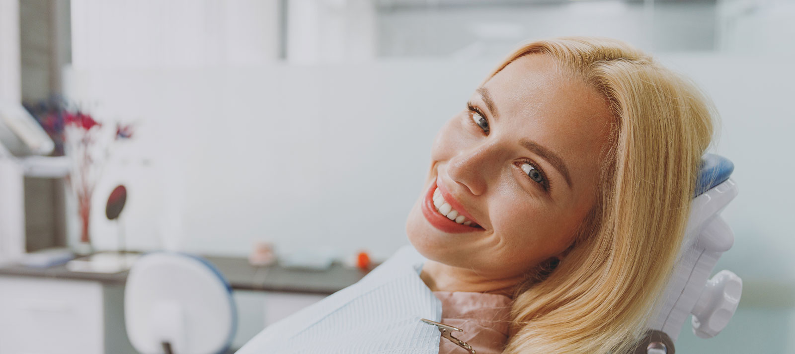 The image features a woman with blonde hair sitting in a dental chair, wearing a red lipstick and smiling at the camera.