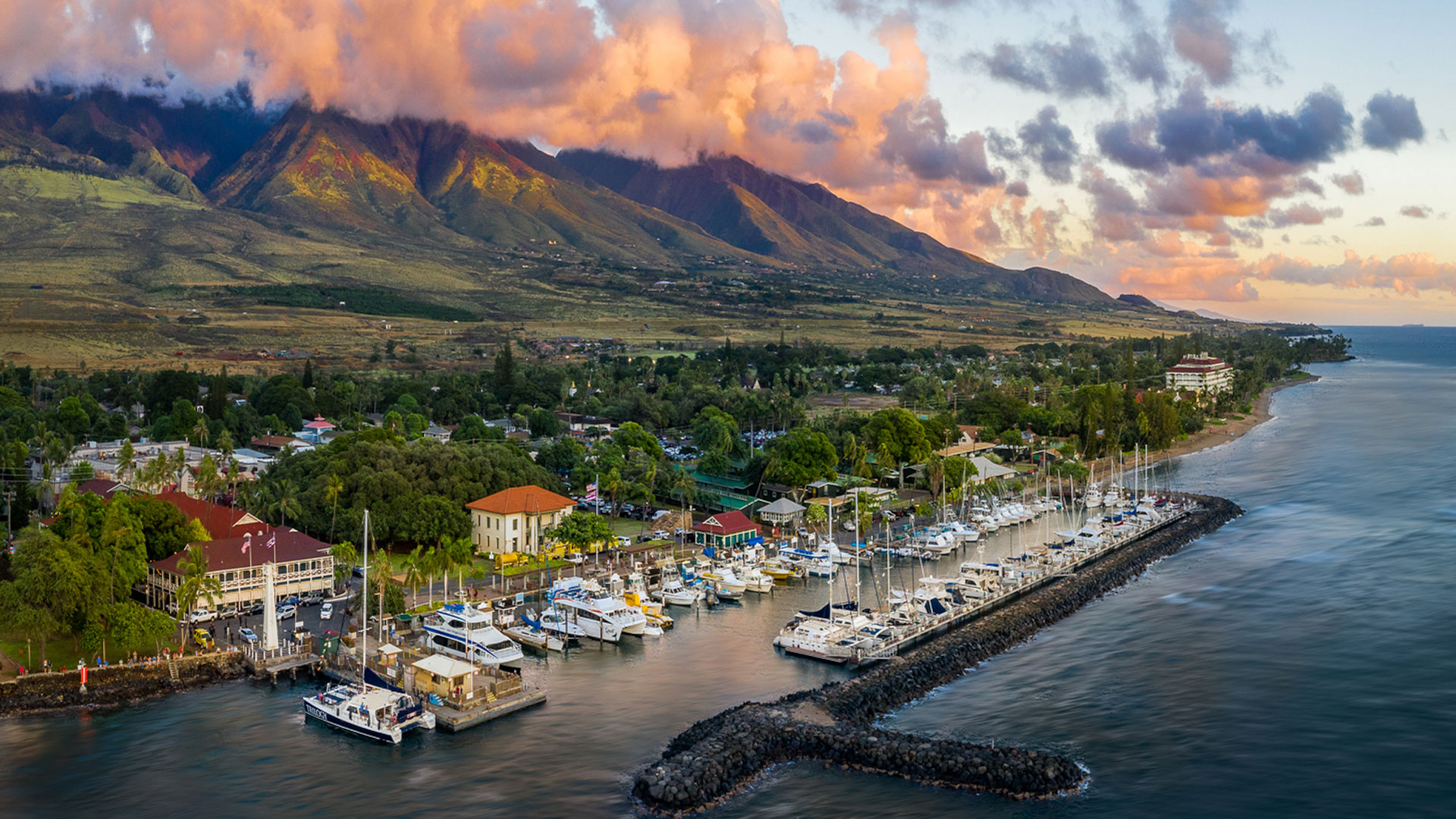 The image depicts a picturesque coastal town with mountains in the background at sunset, featuring a harbor filled with boats, a pier, and colorful buildings along the shoreline.