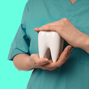 The image shows a dental hygiene professional holding a tooth model with their left hand while wearing blue scrubs.